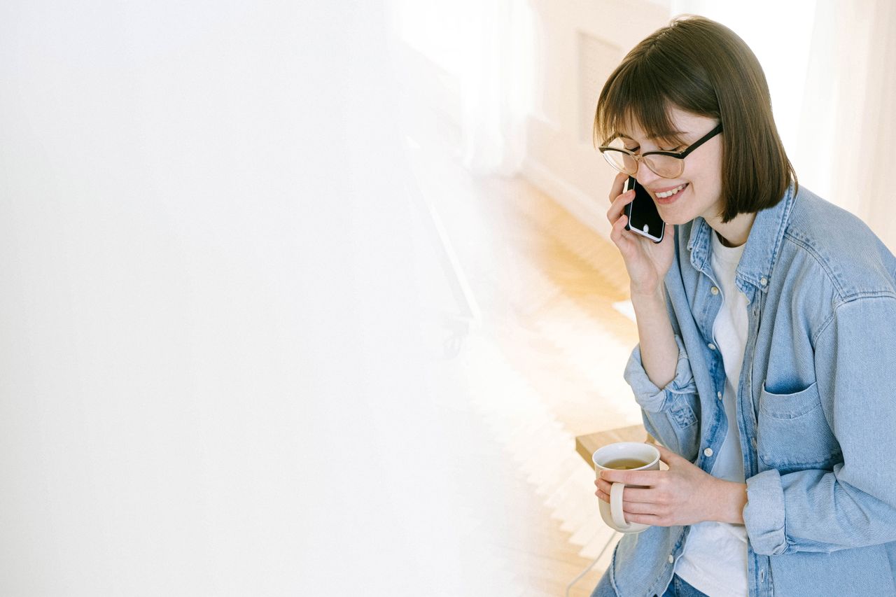 Smiling woman talking on phone with coffee in bright indoor setting.